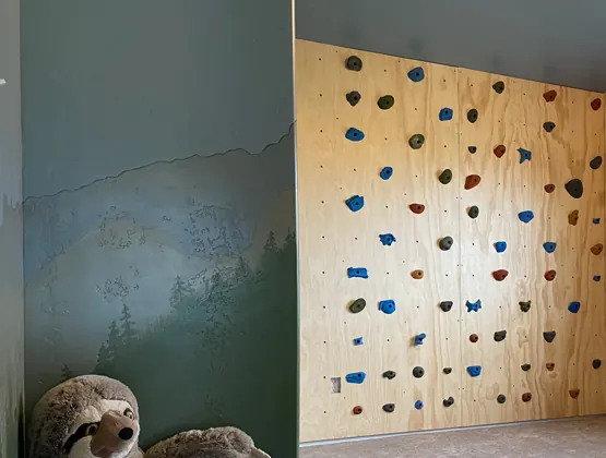 A plywood children’s climbing wall with colorful holds in the corner of a carpeted playroom