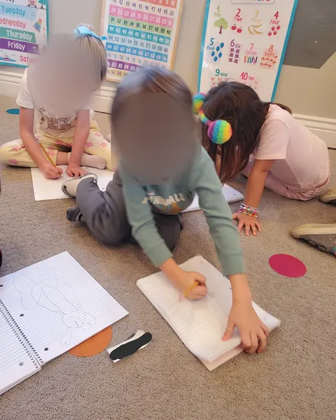 Three preschoolers lying on the classroom floor, writing and drawing on paper