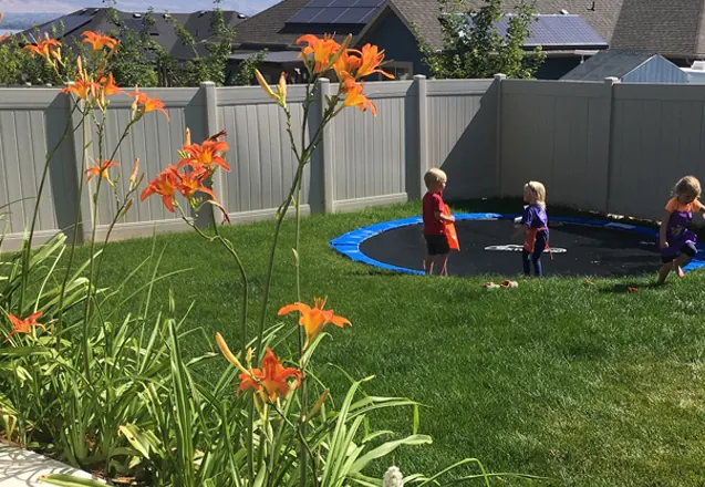 Three kids playing on an in-ground trampoline in a fenced backyard lined with orange lilies